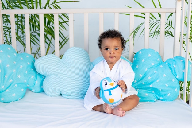 An African baby in a white robe after a bath and bathing sits in bed at home before going to bed and plays with a toy a concept of care and hygiene for young children