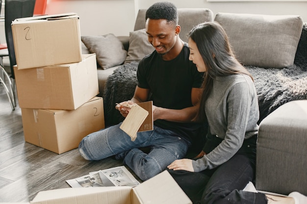 African boy and Asian girl are sitting on a floor in the living room near the boxes with their things. Couple is thinking about a home furnishing