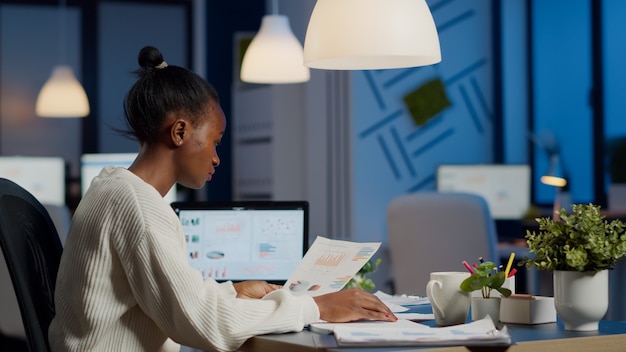 African business woman holding documents with graphics and financial statistics working overtime in front of laptop sitting in start-up business office. Focused employee respecting deadline of project