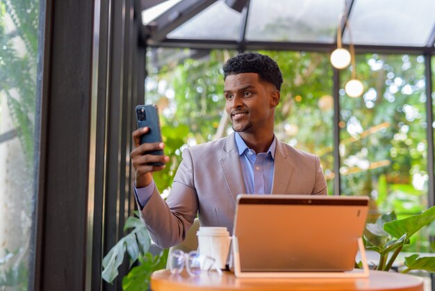 African businessman at coffee shop using laptop computer and taking selfie with mobile phone