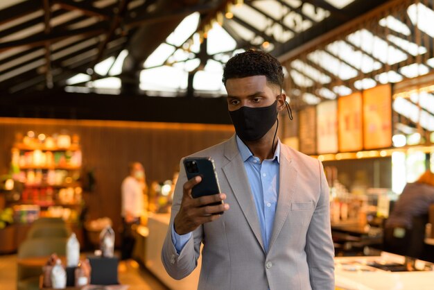 African businessman at coffee shop wearing face mask and using mobile phone