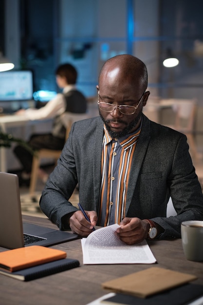 안경을 쓴 아프리카 사업가는 직장에 앉아서 늦은 저녁까지 사무실에서 일하는 동안 문서에 서명합니다.