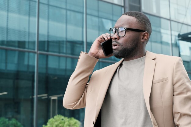 African businessman in eyeglasses talking on his mobile phone while walking along the street in the city