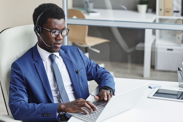 Imprenditore africano in cuffie digitando sulla tastiera del computer al suo posto di lavoro in ufficio