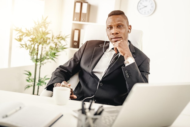 African businessman is sitting in the office, drinking coffee and listening  music in headphones.