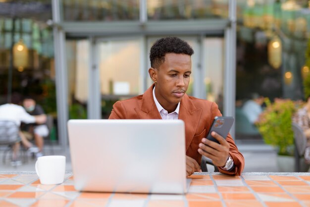 African businessman outdoors in coffee shop using laptop computer and mobile phone