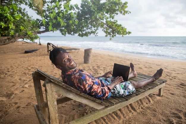 African businessman relaxes at a tropical beach on vacation while using his laptop lying on a sunbed