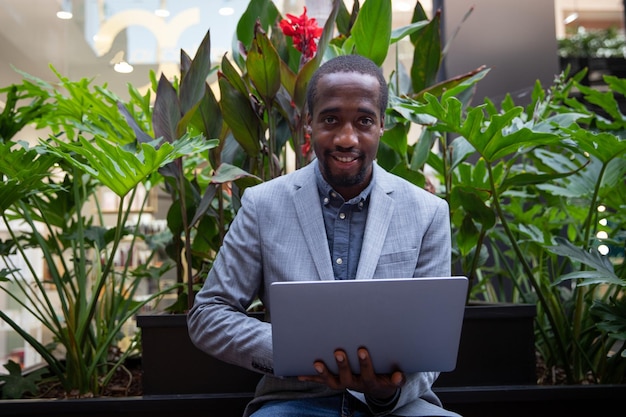 벤치에 앉아 있는 아프리카 사업가가 노트북을 가지고 일합니다. 자신감 있는 표정으로 성공한 사람은 미소를 짓습니다. 사업가 야외에서 작동
