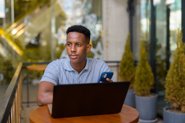 African businessman sitting at coffee shop while using laptop computer and mobile phone while thinking