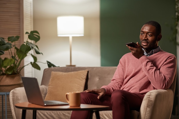 African businessman sitting on sofa in front of table with laptop and recording audio message on his mobile phone he working at home