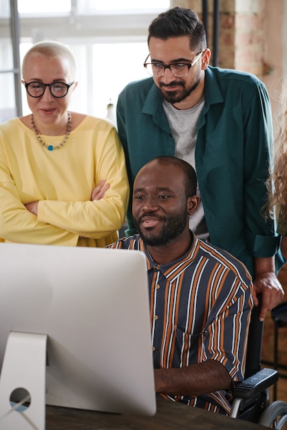 African businessman sitting at the table in front of computer and showing online presentation to his colleagues at office