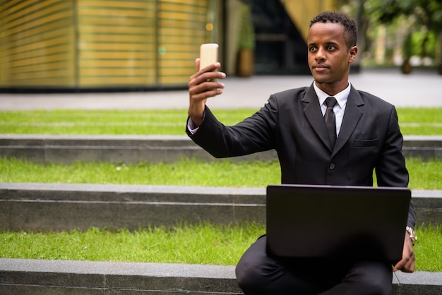 Uomo d'affari africano seduto e utilizzando computer portatile e telefono cellulare all'aperto