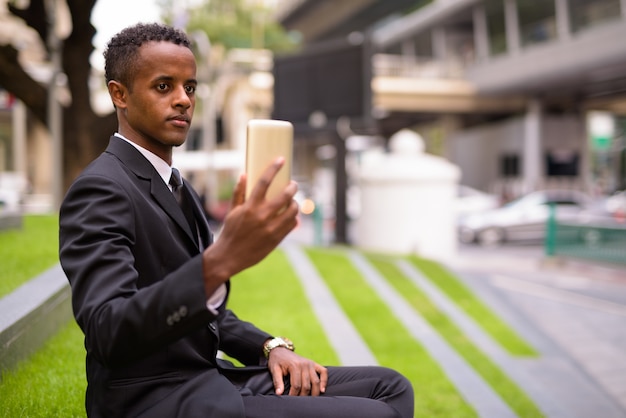 Uomo d'affari africano seduto e utilizzando il telefono cellulare all'aperto