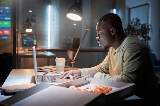 Uomo d'affari africano seduto sul posto di lavoro e facendo uno spuntino con pizza e caffè mentre lavora al computer portatile in ufficio fino a tarda notte