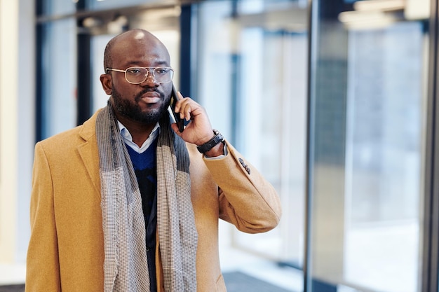 African businessman in stylish coat talking on mobile phone while walking along the office corridor after work