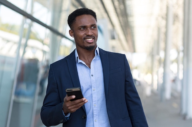 Африканский бизнесмен, путешествующий возле аэропорта с чемоданом, вызывает такси с помощью мобильного приложения