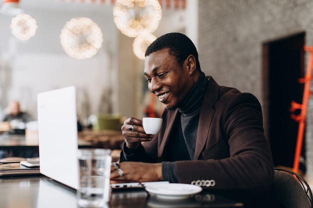 Африканский бизнесмен используя телефон и выпивая кофе пока работающ на компьтер-книжке в ресторане.