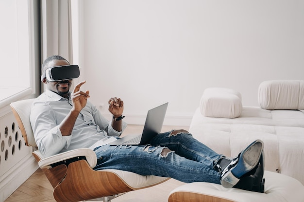 African businessman in virtual reality glasses working at\
laptop sitting in armchair high tech