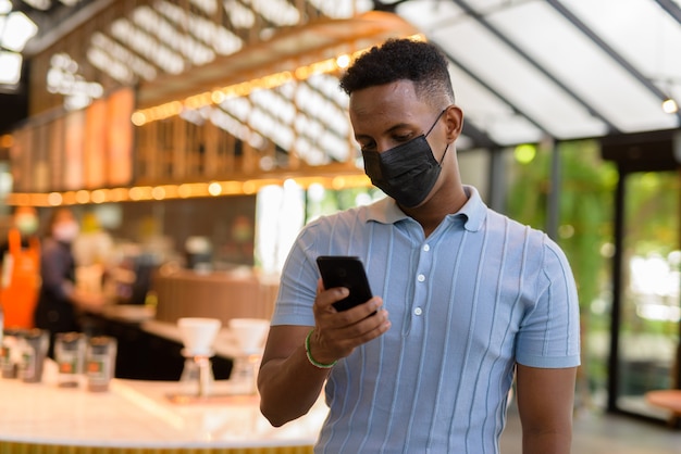 African businessman wearing face mask in coffee shop restaurant while social distancing and using mobile phone