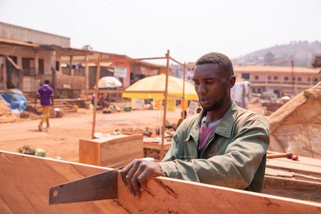 African carpenter in his openair workshop sawing a board tiring\
handcraft