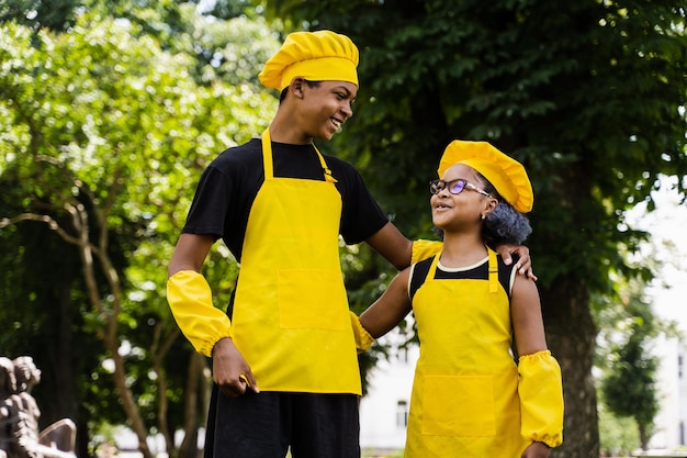 アフリカの子供たちはシェフの帽子と黄色い制服を着て料理をします屋外のアフリカのティーンエイジャーと黒人の女の子は楽しんで料理をします