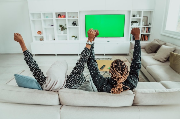 African Couple Sitting On Sofa Watching TV Together Chroma Green Screen Woman Wearing Islamic Hijab Clothes. High quality photo