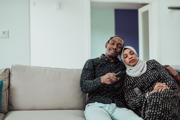 African Couple Sitting On Sofa Watching TV Together. Women Wearing Islamic Hijab Clothes. High-quality photo