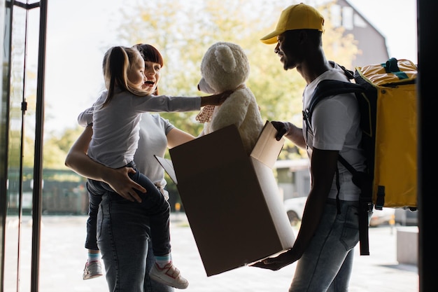 African courier deliver to young attractive female and her daughter a parcel with teddy bear