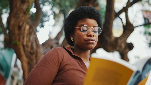 African curlyhaired girl reading a book outdoors wearing glass