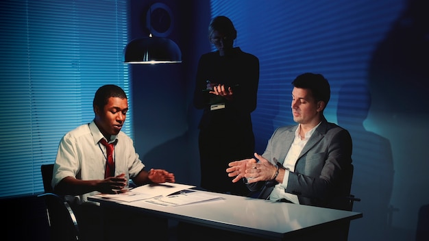 Photo african detective questioning suspect caucasian man in interrogation room female assistant making no...