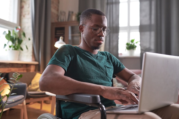 African disabled man sitting in wheelchair with laptop on his knees and typing on it working online at home