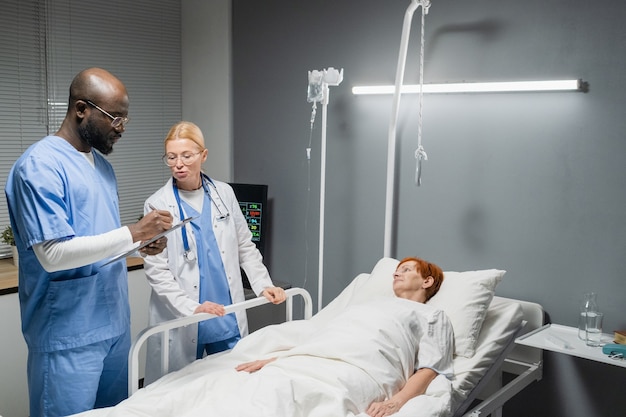 African doctor in uniform writing treatment for senior woman in medical card while standing at hospital ward with nurse