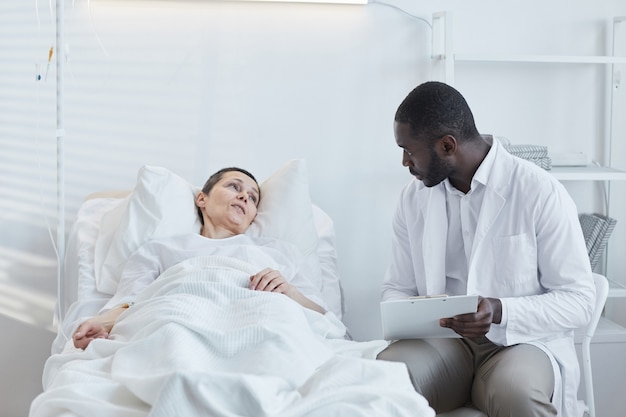 African doctor with medical card talking to patient who lying on hospital bed