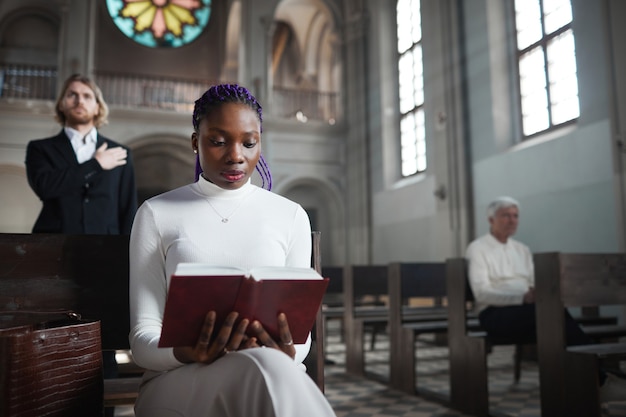 Photo african elegant woman sitting on the bench in the church and reading the bible with other people in the background