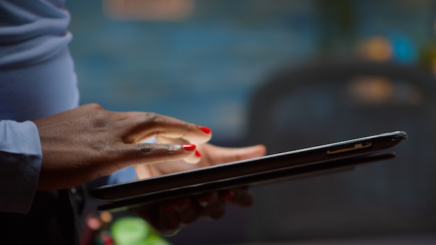 Африканский служащий с помощью планшета, поиск, ввод текста на нем, стоя в гостиной поздно ночью на перерыве. Черный фрилансер, использующий современные технологии беспроводной сети, сверхурочно читает, пишет