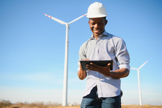 African engineer wearing white hard hat standing with digital tablet against wind turbine on sunny day