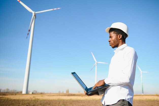 African engineer wearing white hard hat standing with digital tablet against wind turbine