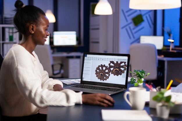Африканский инженер, работающий поздно ночью над 3D-моделью промышленных механизмов за ноутбуком, сверхурочно в стартовом офисе
