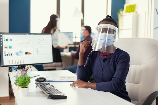 마스크와 얼굴 방패를 쓰고 있는 사무실의 아프리카 기업가는 코로나바이러스를 예방합니다. 글로벌 전염병 동안 사회적 거리를 존중하는 금융 회사에서 일하는 다민족 비즈니스 팀.