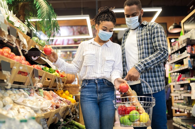 Acquisto della famiglia africana nel supermercato che acquista frutta e verdura insieme in piedi con un cesto pieno di cibo in un negozio di alimentari, indossando maschere per il viso. clienti che scelgono prodotti sani