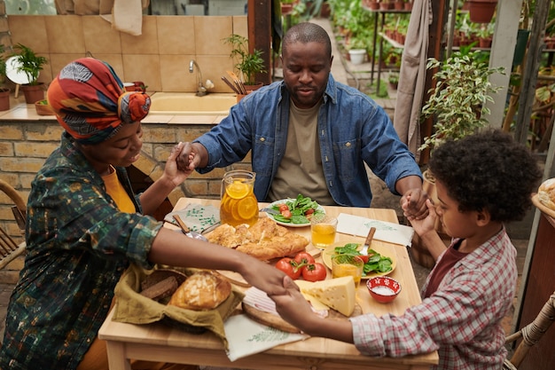 Famiglia africana seduta a tavola e pregando durante la cena in giardino