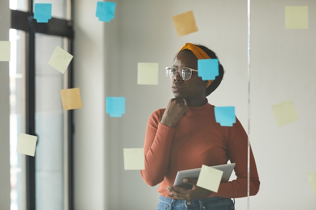 african female designer in eyeglasses standing in front of the glass wall and looking at notes she thinking about new ideas