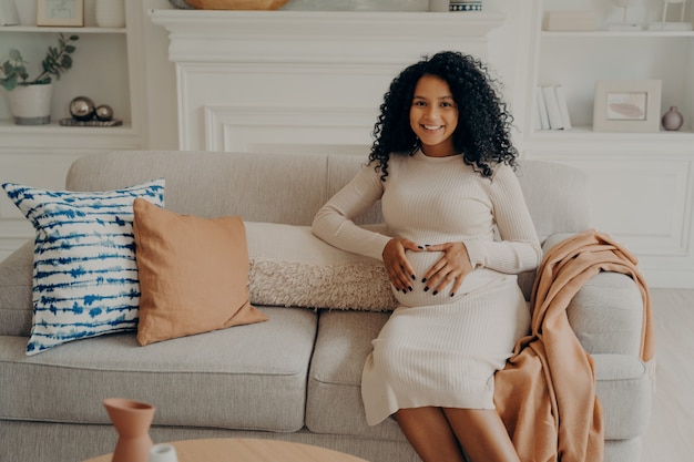 African future mom with bright smile on face keeping hands folded on belly in form of heart