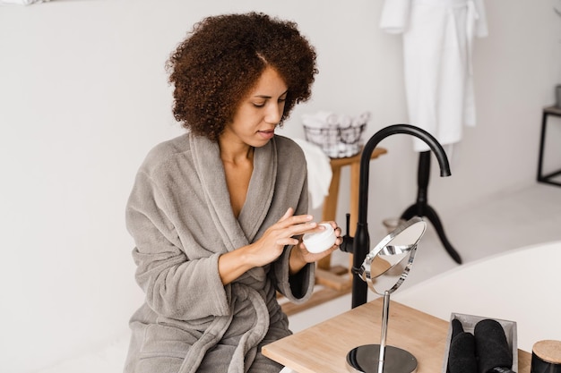 African girl applying face moisturizer cream to protect skin from dryness in the bath African american woman in bathrobe with facial moisturizing cream doing morning beauty routine