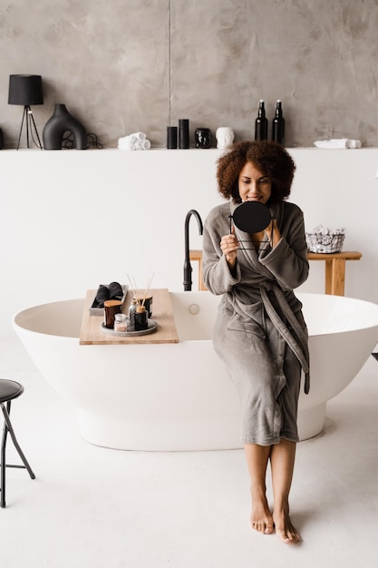 African girl in bathrobe is looking in the mirror on her face in white bathroom African american cheerful woman doing beauty spa procedures and take skin care