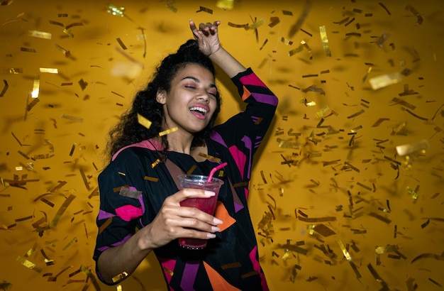 African girl holding drink dancing with confetti isolated on party background