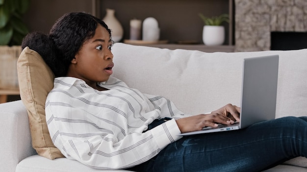 African girl student freelancer user lies on sofa at home looking at laptop using internet checks