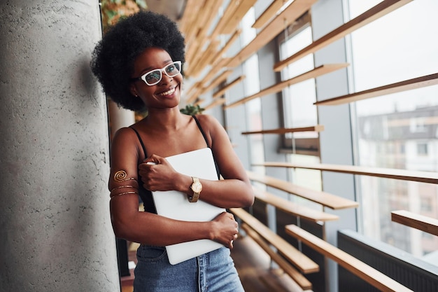 african girl with curly black hair and in casual clothes standing indoors with laptop