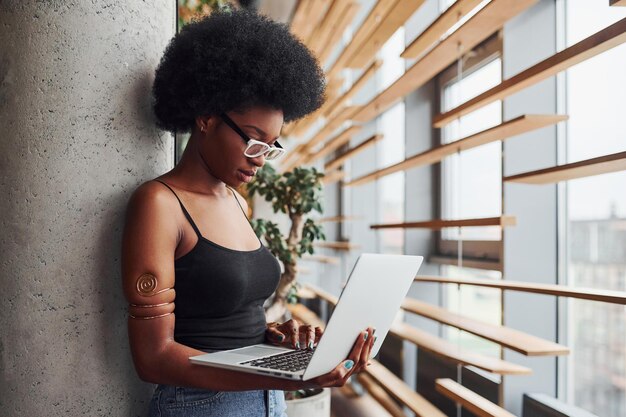 African girl with curly black hair and in casual clothes standing indoors with laptop.