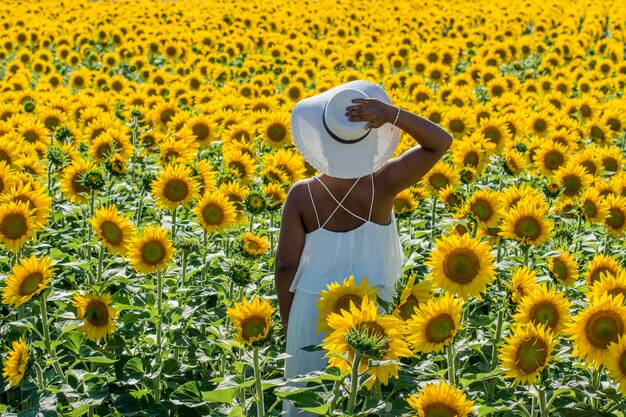 Ragazza africana con abito bianco e cappello sulla schiena nel campo di girasoli durante la giornata di sole